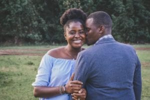 Couple standing in park, woman holding mans arm and smiling at camera, man facing away and kissing her cheek